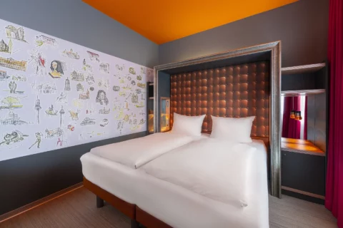 Junior-Suite – Blick diagonal auf Doppelbett. Gerahmtes Poster-Kopfteil in Leder-Optik, eingelassene Nachttische mit Beleuchtung, Hannover-Motivwand links
