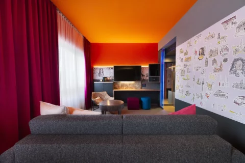 Junior-Suite – Wohnbereich. Sofa Rückseite im Vordergrund, Sitzbereich mit Sessel und Schaukelstuhl und rundem Tisch, Arbeitsplatz mit Hockern und TV/Minibar/Safe in Augenhöhe, Hannover-Motivwand