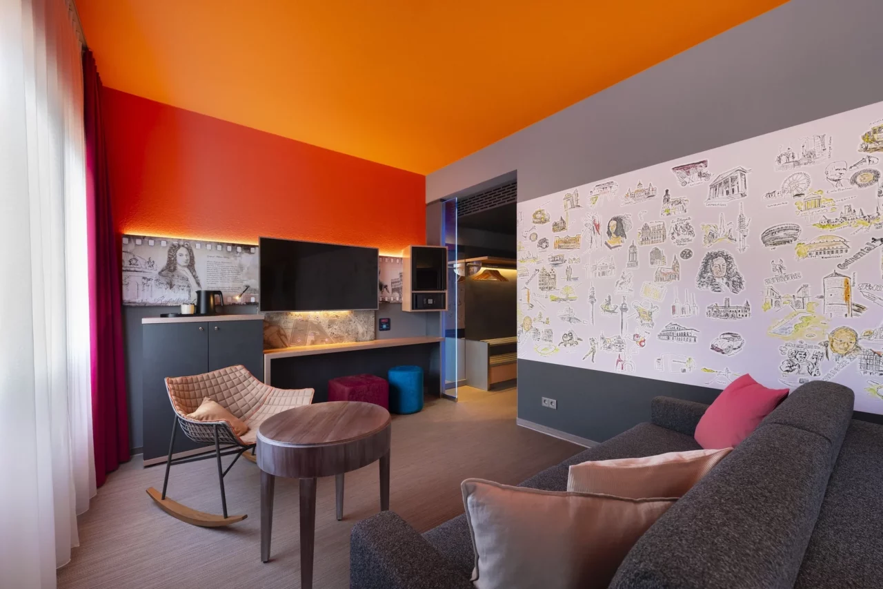 Junior-Suite – Wohnbereich. Sofa mit Kissen links im Vordergrund, Sitzbereich mit Schaukelstuhl und rundem Tisch, Arbeitsplatz mit TV/Minibar/Safe in Augenhöhe, Hannover-Motivwand