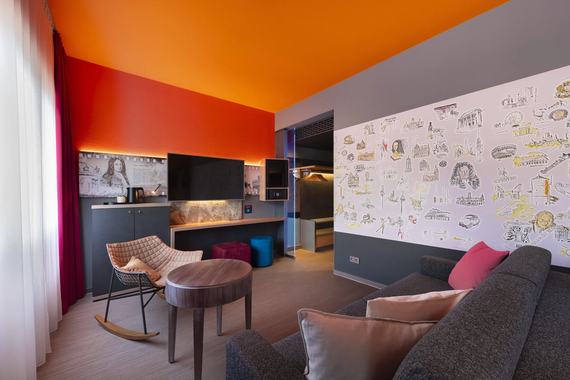 Junior suite - living area. Sofa with cushions on the left in the foreground, seating area with rocking chair and round table, workstation with TV/minibar/safe at eye level, Hanover motif wall