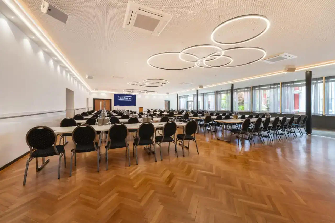 Small hall - angled view from back to front. Parliamentary Seating arrangements with conference equipment, LED display on front wall, full-length window front on the right, modern ring pendant lights, herringbone parquet flooring
