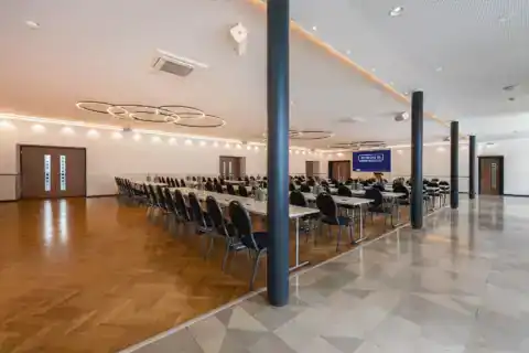 Kleiner Saal – diagonale Ansicht von hinten nach vorne: Parlamentarische Bestuhlung, LED-Display mittig an Stirnwand, moderne Ringpendelleuchten, Fischgrätparkett im Tagungsbereich, vier Säulen als visueller Raumteiler trennen den Bereich mit Fliesenboden