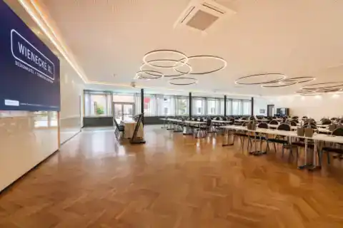 Kleiner Saal – Frontansicht von links. LED-Display an Stirnwand, Rednerpult mittig, parlamentarische Bestuhlung, Fensterfront mit Vorhängen, moderne Ringpendelleuchten, Fischgrätparkett