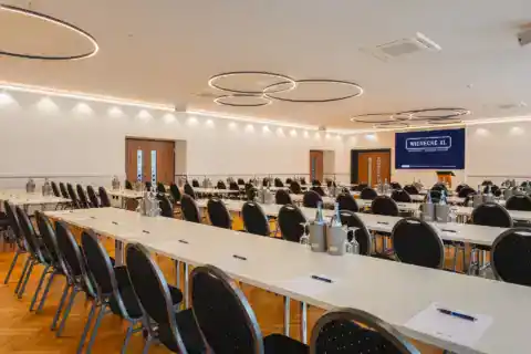Small hall - view from the rows of chairs. Parliamentary Seating arrangements with conference equipment (water bottles, writing pads), LED display on front wall, modern ring pendant lights, herringbone parquet flooring