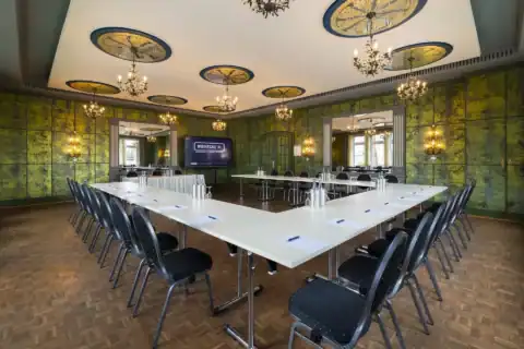 Spiegelzimmer – zentrale Ansicht. U-förmige Bestuhlung, LED-Display mittig an Wand, zwei große ornamentale Wandspiegel links und rechts, neun ornamentale Deckenrosetten mit floralen Kronleuchtern, historische grüne Wandgestaltung mit ornamentalen Kerzen-Optik-Wandleuchten, Parkettboden