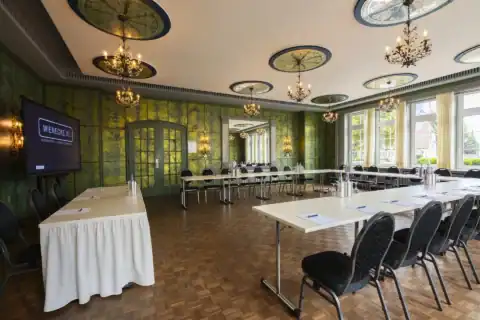 Spiegelzimmer – seitliche Ansicht mit Wandspiegel. U-förmige Bestuhlung mit Präsidiumstisch, großer ornamentaler Wandspiegel mittig, LED-Display links, neun ornamentale Deckenrosetten mit floralen Kronleuchtern, historische grüne Wandgestaltung mit Wandleuchten, Fensterfront rechts, Parkettboden