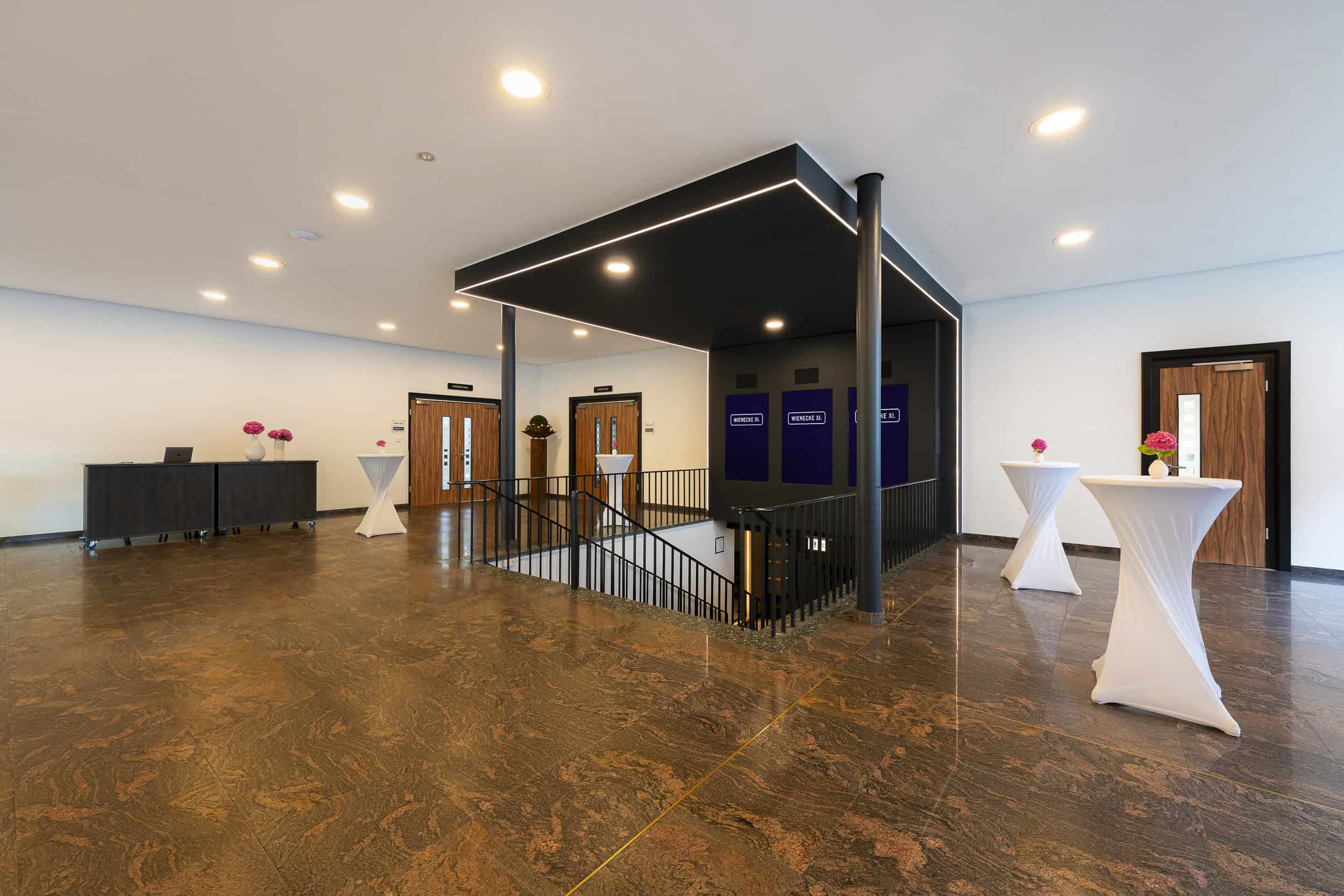 Ansicht Foyer Kleiner Saal mit weißen Stehtischen neben der Treppe, links zwei fahrbare dunkle Tische mit Blumen und Laptop, dunkle marmorierte Fliesen, helle Deckenspots, absteigende Treppe, drei LED-Displays über der Treppe