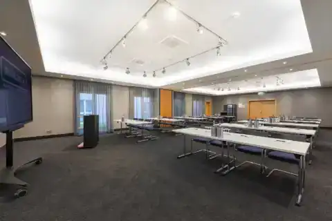 Tagungsraum Hotel – diagonale Ansicht von vorne zum Eingang. Parlamentarische Bestuhlung mit blauen Freischwingern, LED-Display links an Wand, Rednerpult vorne, zwei eingelassene Deckenbereiche mit indirekter Beleuchtung und Schienenstrahler-System, bodentiefe Fenster, grauer Teppichboden
