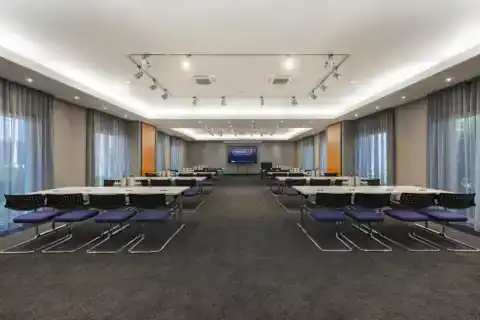 Tagungsraum Hotel – zentrale Ansicht nach vorne. Parlamentarische Bestuhlung für 40 Personen mit blauen Freischwingern, LED-Display mittig an Stirnwand, zwei eingelassene Deckenbereiche mit indirekter Beleuchtung und Schienenstrahler-System, bodentiefe Fenster beidseitig mit Vorhängen, grauer Teppichboden