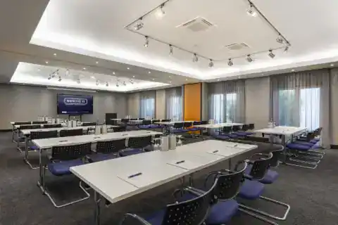Conference room hotel - diagonal view to the front. Parliamentary Seating arrangements for 40 people, with blue cantilever chairs, LED display on front wall, two recessed ceiling areas with indirect lighting and adjustable spotlights, floor-to-ceiling windows on the right with curtains, carpeting