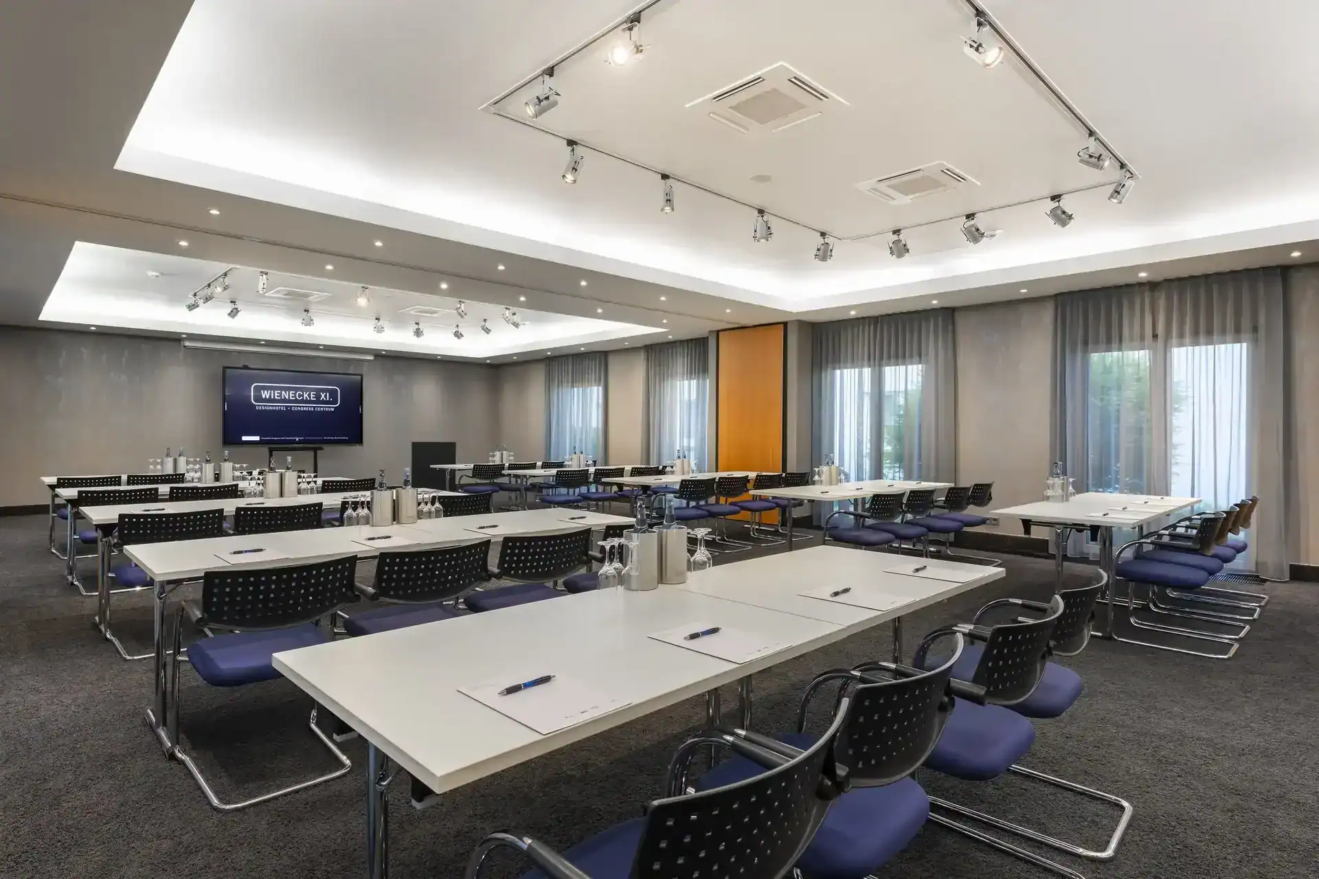 Tagungsraum Hotel – diagonale Ansicht nach vorne. Parlamentarische Bestuhlung für 40 Personen, mit blauen Freischwingern, LED-Display an Stirnwand, zwei eingelassene Deckenbereiche mit indirekter Beleuchtung und verstellbaren Strahlern, bodentiefe Fenster rechts mit Vorhängen, Teppichboden