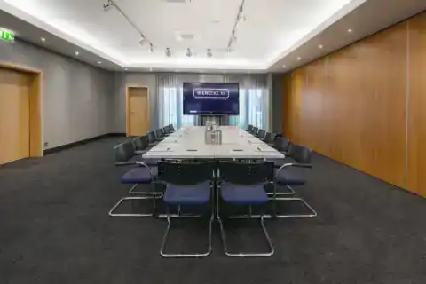 agungsraum I – Frontalperspektive. Konferenztisch mit Freischwingern, Display zwischen den Fenstern, Wand mit Tür links, Holzvertäfelung rechts, Deckenstrahler und indirekte Beleuchtung