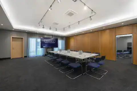 Tagungsraum I – Diagonale Perspektive. Konferenztisch mit Freischwingern, mobiles Display zwischen den Fenstern, Holzvertäfelung mit geöffneter Verbindungstür zum Nachbarraum, Deckenstrahler