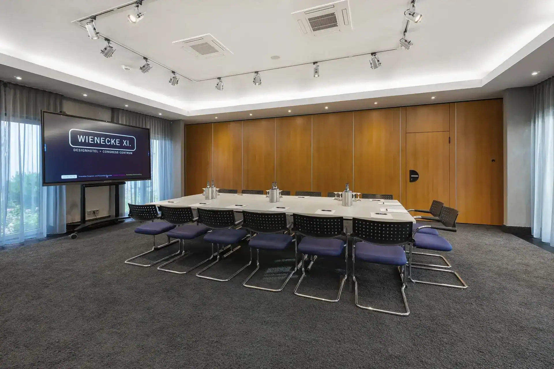 Tagungsraum I – Rechteckiger Konferenztisch mit Freischwinger-Stühlen, mobiles Display, Holzvertäfelung an einer Seite, Deckenstrahler und indirekte Beleuchtung, Teppichboden