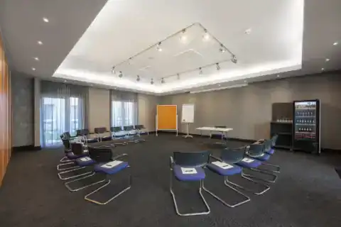 Tagungsraum II – Stuhlkreis-Bestuhlung mit Freischwingern und einem Tisch, Flipchart und Pinnwand, Self-Service Getränkekühlschrank, zwei bodentiefe Fenster, verschiedene Deckenstrahler