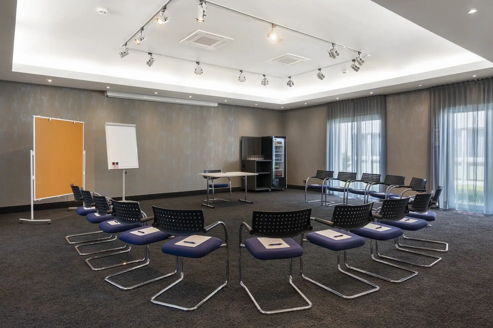 Tagungsraum II – Diagonale Perspektive: Im Zentrum Stuhlkreis-Bestuhlung, mit Freischwingern und einem Tisch. Self-Service Getränkekühlschrank in der gegenüberliegenden Raumecke. Flipchart und Pinnwand links, zwei bodentiefe Fenster rechts, verschiedene Deckenstrahler