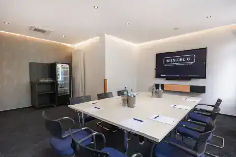 Conference room V - Conference table with 8 cantilever chairs, display on the wall opposite, drinks refrigerator on the left, indirect lighting, carpeting