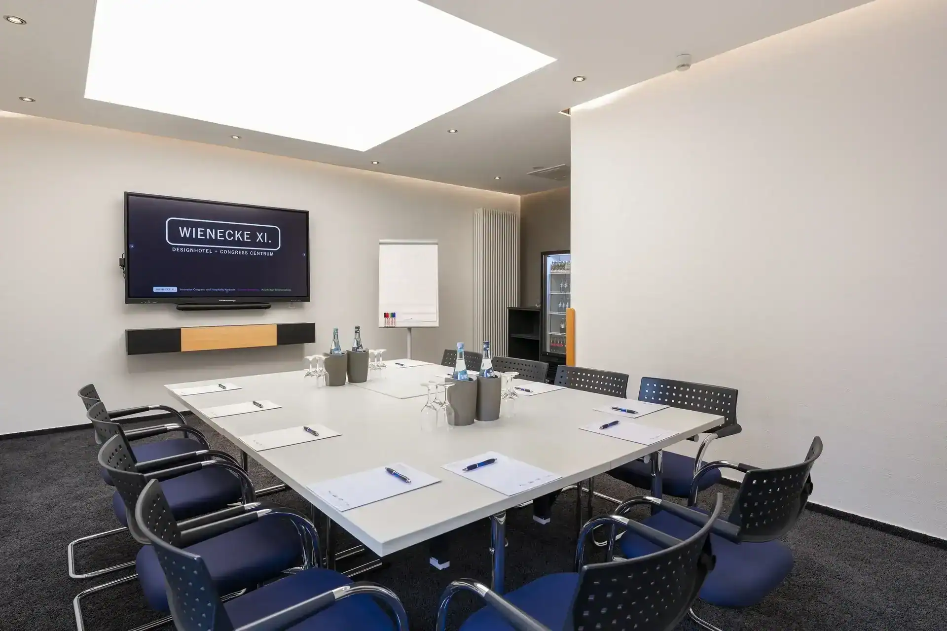 Tagungsraum VI – Perspektive Konferenztisch zu Wanddisplay. Konferenztisch mit 10 Freischwingern, Display an Stirnwand, Flipchart, Getränkekühlschrank, indirekte Deckenbeleuchtung, Oberlicht