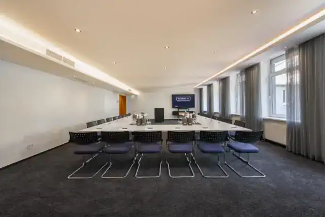 Tagungsraum VII – Frontalperspektive. U-Form Bestuhlung mit Konferenztischen und 24 Freischwingern, Display an Stirnwand, Rednerpult, Fensterfront rechts, indirekte Deckenbeleuchtung, Teppichboden