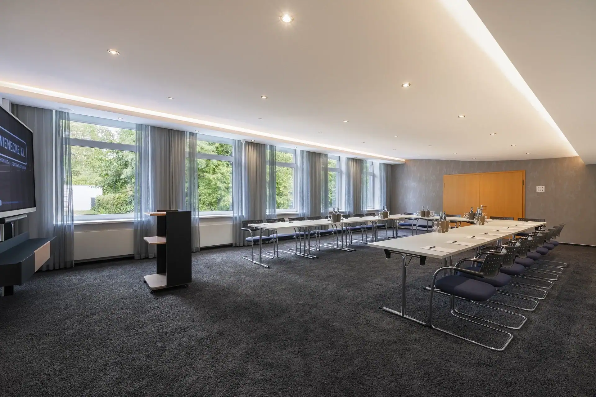 Tagungsraum VII – Blick Rückwand zu Eingangstür aus Holz. U-Form Bestuhlung mit Konferenztischen und 24 Freischwingern, Display links, Rednerpult mit Stauraum, Fensterfront, Teppichboden