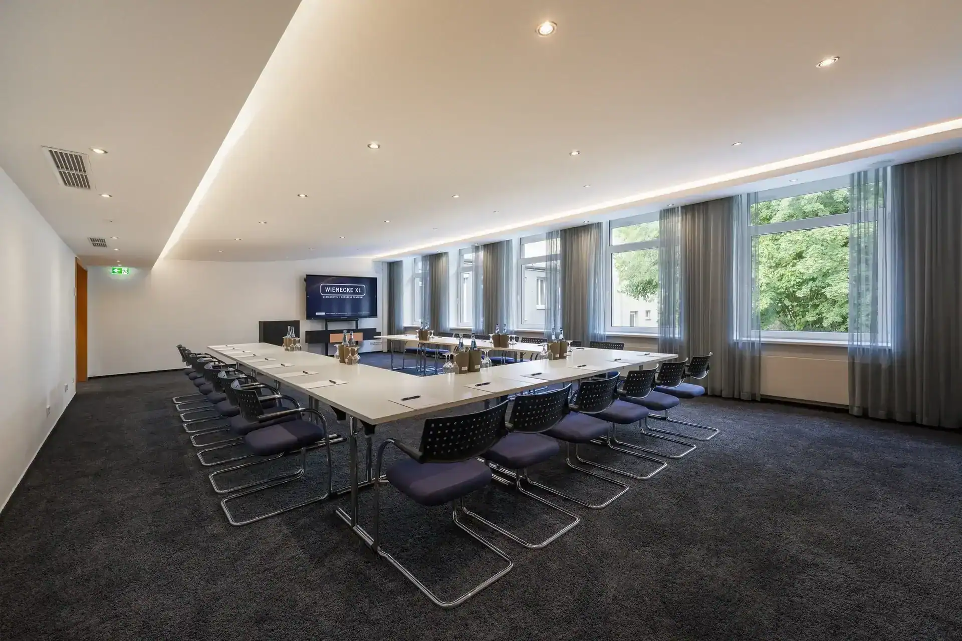 Tagungsraum VII – Diagonale Perspektive. U-Form Bestuhlung mit Konferenztischen und 24 Freischwingern, Rednerpult, mobiles Display, Fensterfront (5 Fenster), indirekte Deckenbeleuchtung, Teppichboden