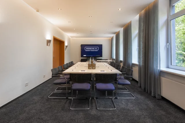 Tagungsraum VIII – Frontalperspektive. Konferenztisch mit 14 Freischwingern, Display an Stirnwand, Verbindungstür links, Fensterfront rechts, Wandleuchten und Einbauspots, Teppichboden