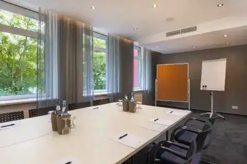 Tagungsraum VIII Detail – Plätze am Konferenztisch mit Schreibausstattung und Getränken, Pinnwand und Flipchart dahinter, Fensterfront links, Teppichboden