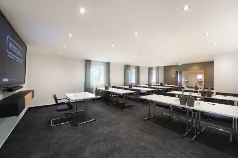 Tagungsraum XI – Diagonale Rückansicht. Parlamentarische Bestuhlung mit Tischen und 25 Freischwingern, Display links, drei Fenster, offene Verbindung zum Vorraum rechts, Einbauspots, Teppichboden