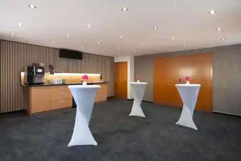 Tagungsraum XI Vorraum – Drei Stehtische mit Hussen, Kaffeestation mit Kaffeevollautomat und Geschirr, Holzlamellenwand links, Doppeltür rechts, Einbauspots, Teppichboden