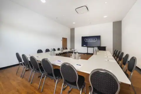 Tagungsraum XX – Frontalperspektive. U-Form Bestuhlung mit 18 Polsterstühlen, Display an Stirnwand, Tür links davon, Holzboden, Einbauspots