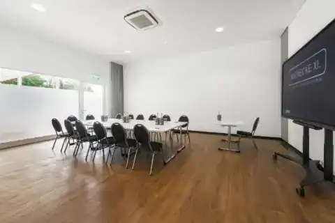 Conference room XXI - Diagonal perspective. Rectangular table arrangement with 15 upholstered chairs, mobile display on the right, lectern, wooden floor, floor-to-ceiling windows on the left with privacy film