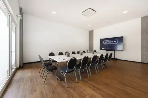 Tagungsraum XXI – Diagonale Perspektive. Geschlossene Tischanordnung mit 15 Polsterstühlen, Display an der Wand, bodentiefe Fenster links, Holzboden, Einbauspots