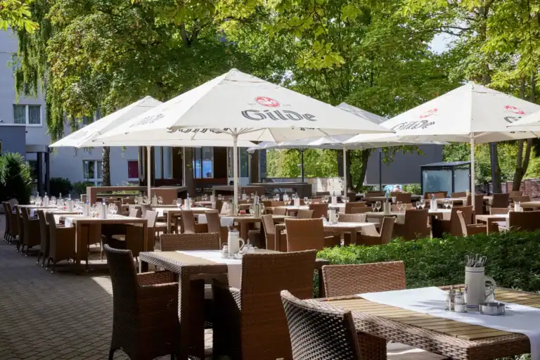 Sonnige Terrasse mit 20 Tischen für je 3-4 Personen. Außen-Mobiliar in geflochtenem Stil. Große Sonnenschirme, rechts grüne Hecke, im Hintergrund mit Bäumen umgeben