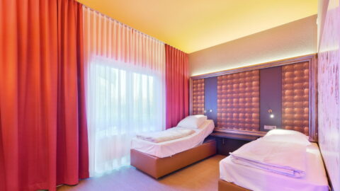 Behindertengerechtes Zimmer – zwei elektrisch verstellbare Einzelbetten mit viel Abstand. Regal als Nachttisch zwischen den Betten und wandmontierte, schwenkbare Lampen.