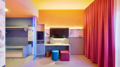 Behindertengerechtes Zimmer – Arbeitsbereich und Garderobe. Offene Garderobe links, unterfahrbarer Schreibtisch mit TV darüber, Minibar und Safe in Regal mit Stauraum, im Sitzen erreichbar, zwei Hocker