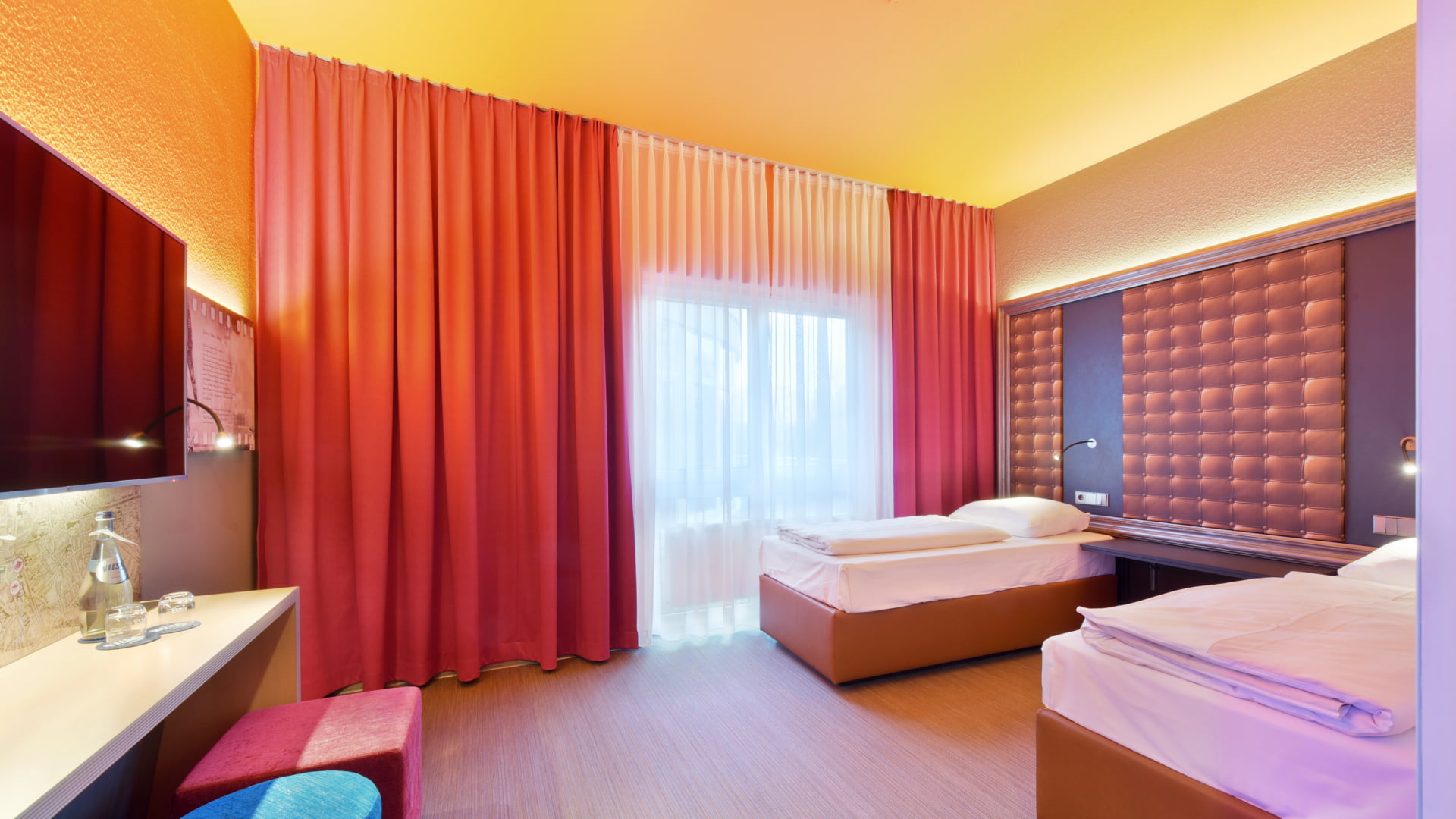 Behindertengerechtes Zimmer Ansicht mit zwei getrennten Einzelbetten. Ein transparenter und roter Vorhang mit Tageslicht. Links ist ein TV, Schreibtisch und zwei moderne Hocker in Rot und Violett.