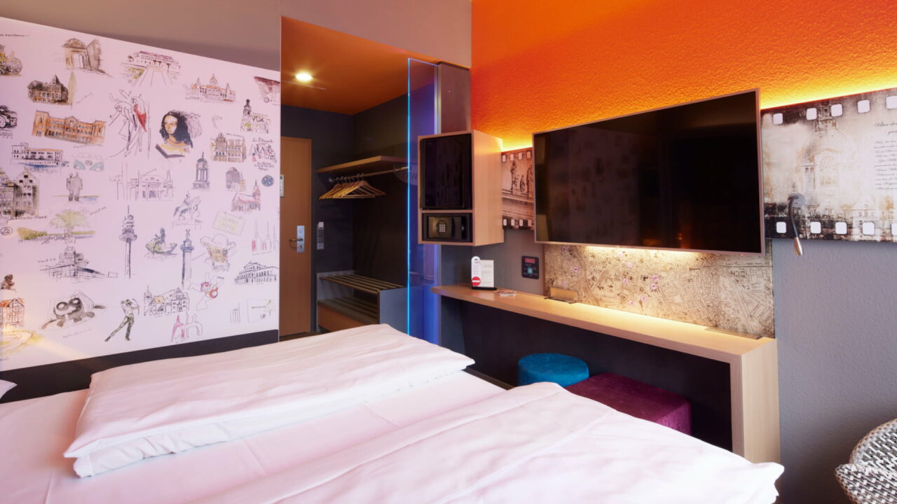 Zweibettzimmer – Perspektive vom Bett zum Eingang. Hannover-Motivwand, Arbeitsplatz, darüber TV, Mini-Kühlschrank und Safe in Augenhöhe, offene Garderobe