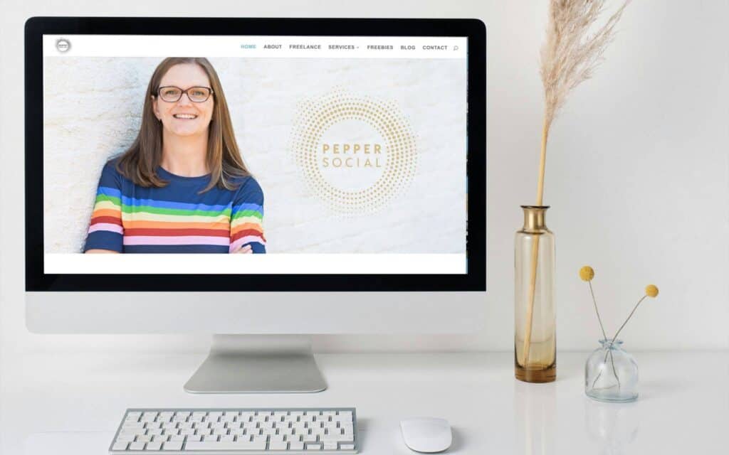 A computer on a white desk displays a small business website featuring a person in a colorful striped shirt and glasses smiling. The website header reads Pepper Social. Beside the monitor is a vase with tall, dried grass, while a keyboard and mouse sit in front. Wildcurrant Marketing