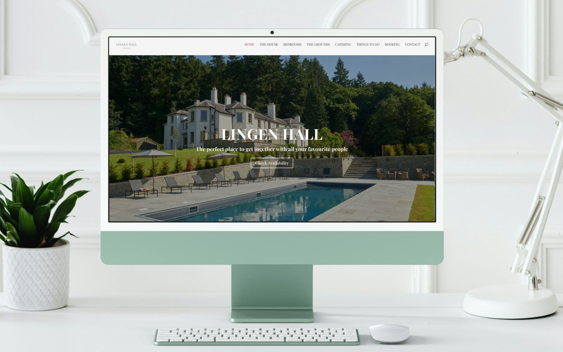 A computer screen showcases a luxurious property website with impeccable web design, featuring a grand mansion and pool. Lush greenery enhances the scene. A white keyboard, mouse, plant, and desk lamp complete the setup, ensuring both elegance and SEO optimization. Wildcurrant Marketing