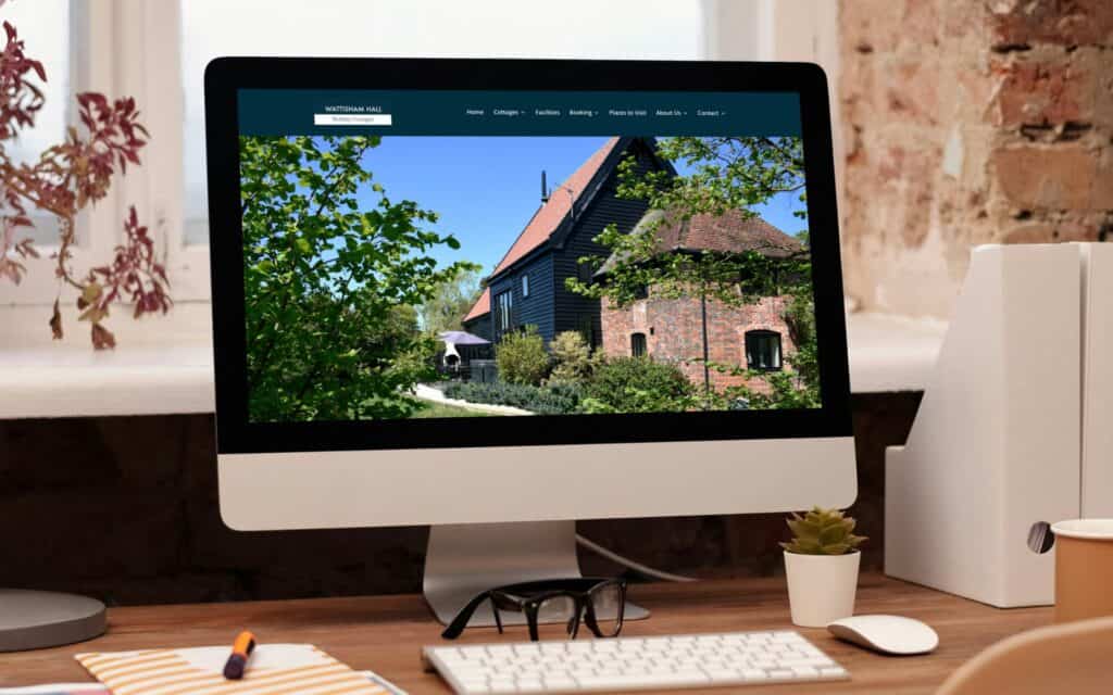 A computer monitor on a wooden desk showcases an image of a rustic brick and wooden house, reflecting a web design with SEO focus. Potted plants, a notebook, and a pencil accompany the scene. Natural light streams through the window, enhancing the workspace’s charm. Wildcurrant Marketing