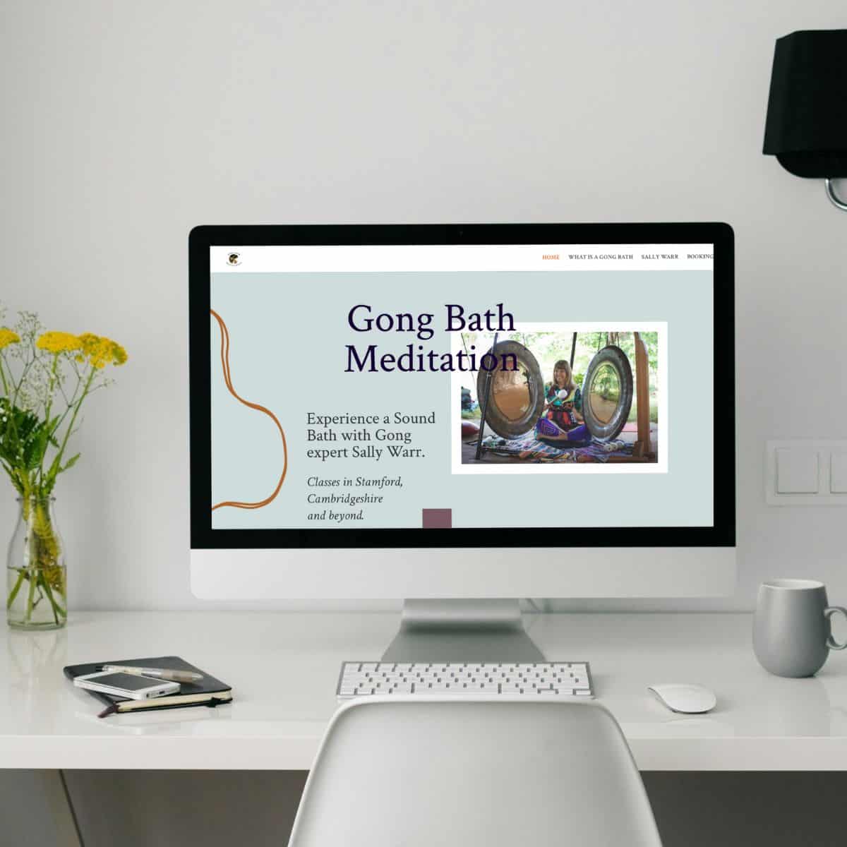 A computer screen displays a website for Gong Bath Meditation with an SEO-friendly design, featuring an image of a person playing gongs. The desk holds a plant, notepad, cup, and keyboard. The background is light and minimalistic, emphasizing the importance of digital harmony. Wildcurrant Marketing