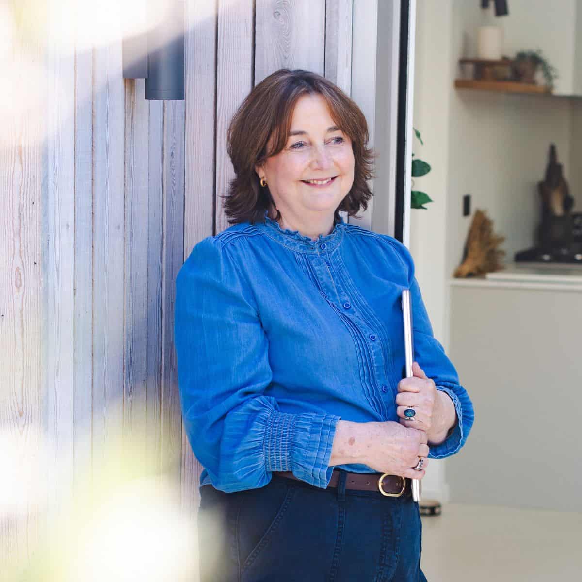 A woman with shoulder-length brown hair, wearing a blue blouse and dark pants, stands by a wooden wall holding a closed laptop and smiling. The background shows a modern, light-filled room. Wildcurrant Marketing