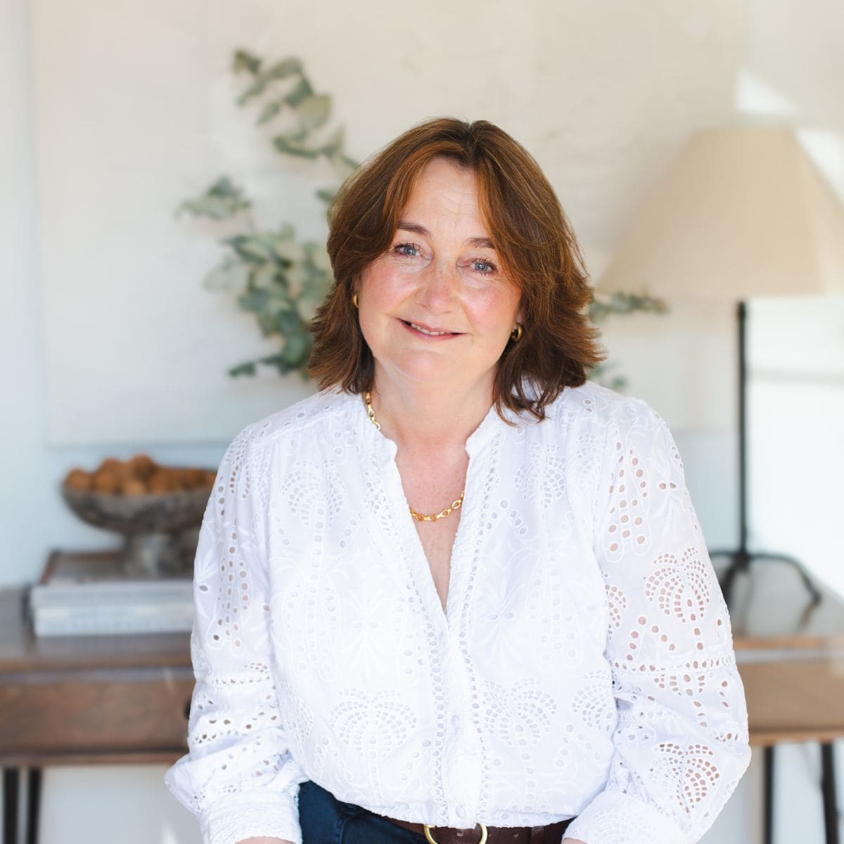 A woman with shoulder-length brown hair, wearing a white eyelet blouse and gold jewelry, sits indoors in a softly lit room with a table, books, and a lamp in the background. She is smiling gently at the camera. Wildcurrant Marketing