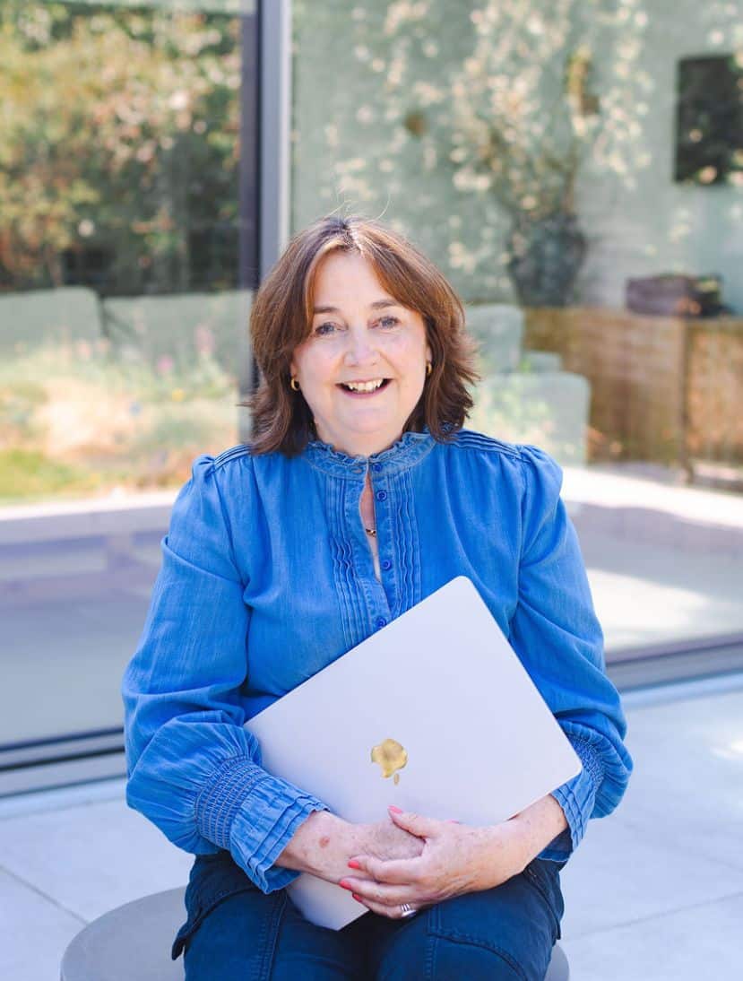 About: A woman with brown hair wearing a blue blouse sits outdoors, smiling and holding a closed silver laptop with a gold apple sticker on it. Lush greenery and a glass window set the scene in the background. Wildcurrant Marketing