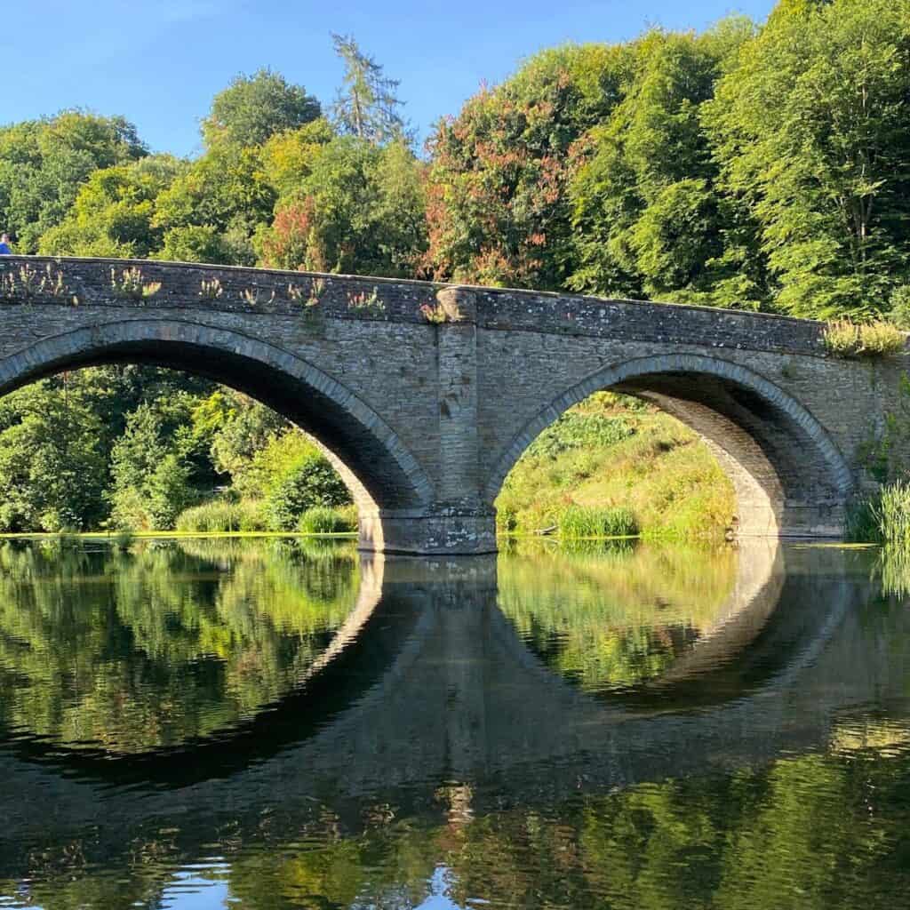 A stone bridge with two arches spans over a calm river, reflecting perfectly in the water. Lush green trees surround the area beneath a clear blue sky—much like how WEB DESIGN HEREFORD brings natural harmony to digital landscapes. Wildcurrant Marketing