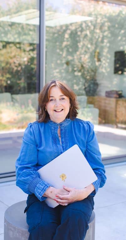 Smiling woman in a blue blouse holding a laptop, sitting outside with a modern glass building in the background, promoting confidence and success.