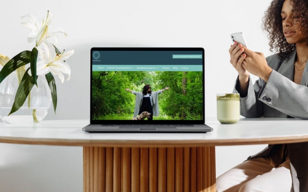 A woman using a laptop with a website on the screen, sitting at a white table with a flower vase.
