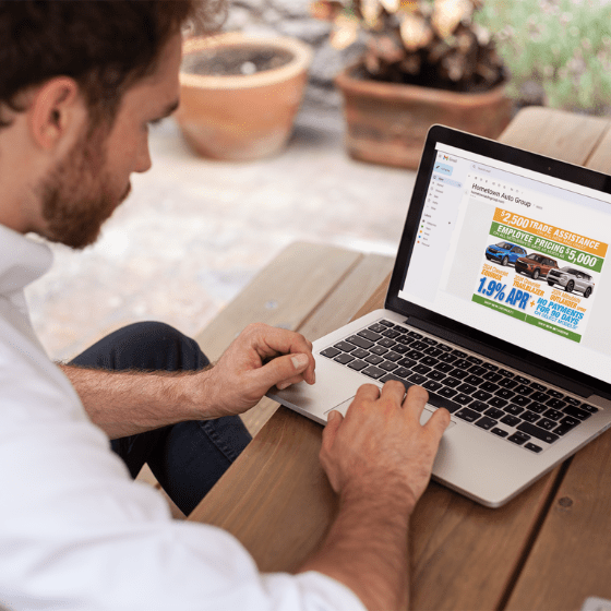 Man at wooden table using laptop displaying car dealership ad featuring vehicle images and promotional offers, including trade-in assistance and low APR financing—targeted marketing for Willowood Ventures automotive sales.