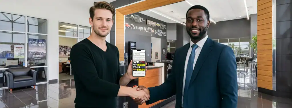 Two men shake hands in a modern car dealership showroom; one holds a phone showing “We need your trade” on the screen. Cars and large windows are visible in the background, highlighting vehicle trade-in opportunities at Willowood Ventures.