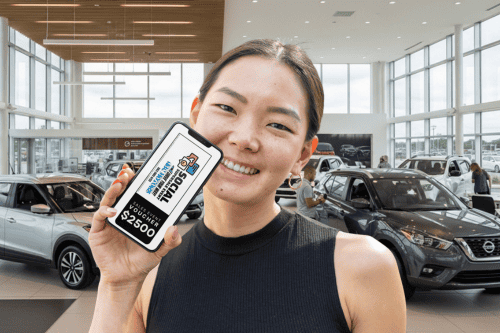 A woman in a car dealership showroom holds up a smartphone showing a $2,500 voucher for a social media giveaway, highlighting effective automotive marketing by Willowood Ventures. New cars and customers are visible in the bright dealership background.